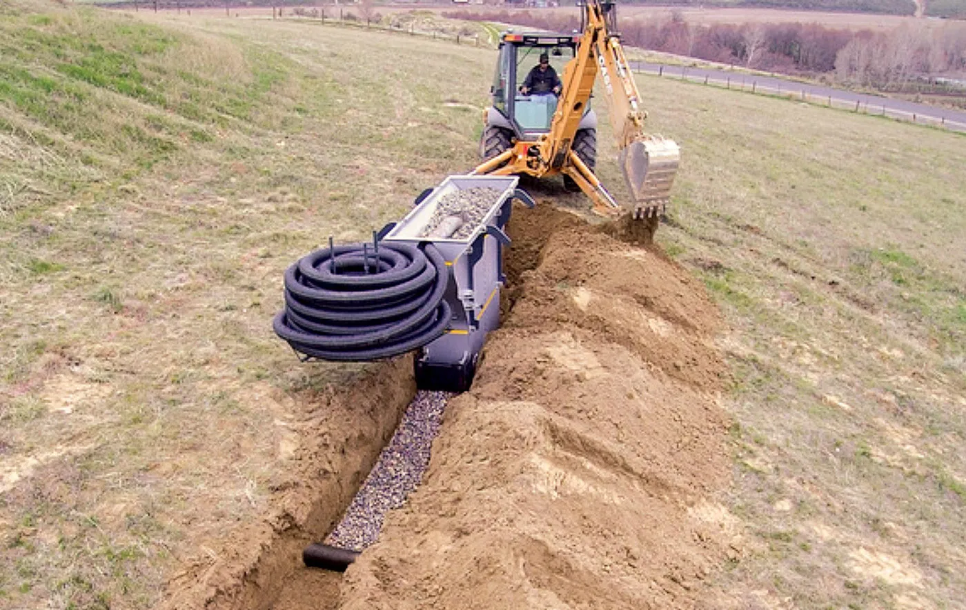 Backhoe filling a narrow drain trench with clean stone beside a coil of black perforated pipe