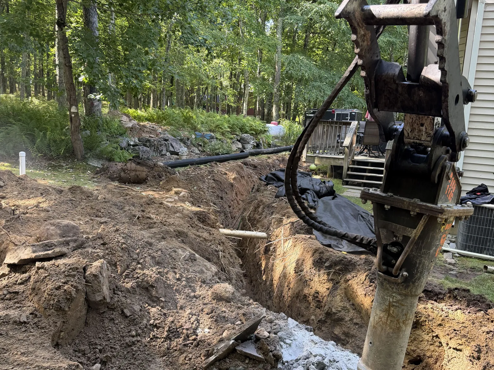 Hydraulic breaker working ledge at the bottom of a trench on a wooded lot