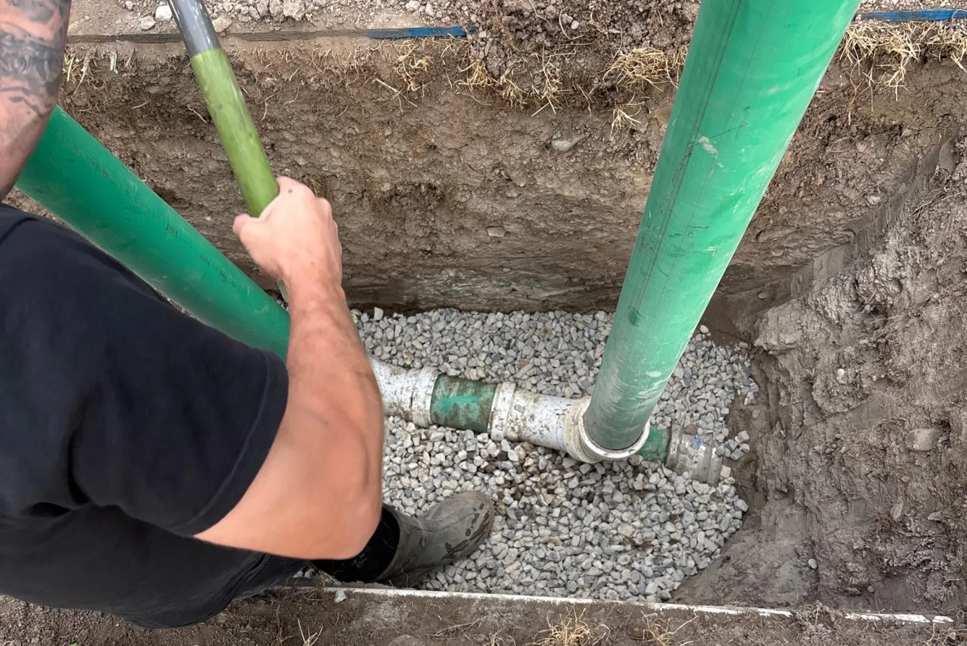 Worker joining green PVC sewer pipe over a stone bed in an open residential trench
